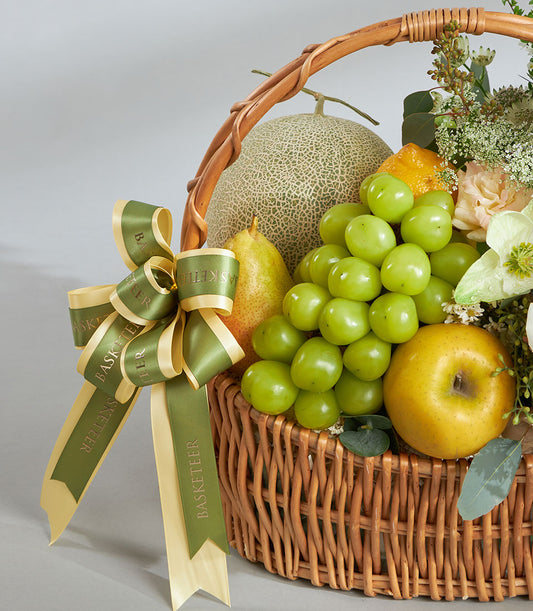 Premium green and white fruit basket with imported cantaloupe, grapes, pear, and fresh flowers in woven wicker