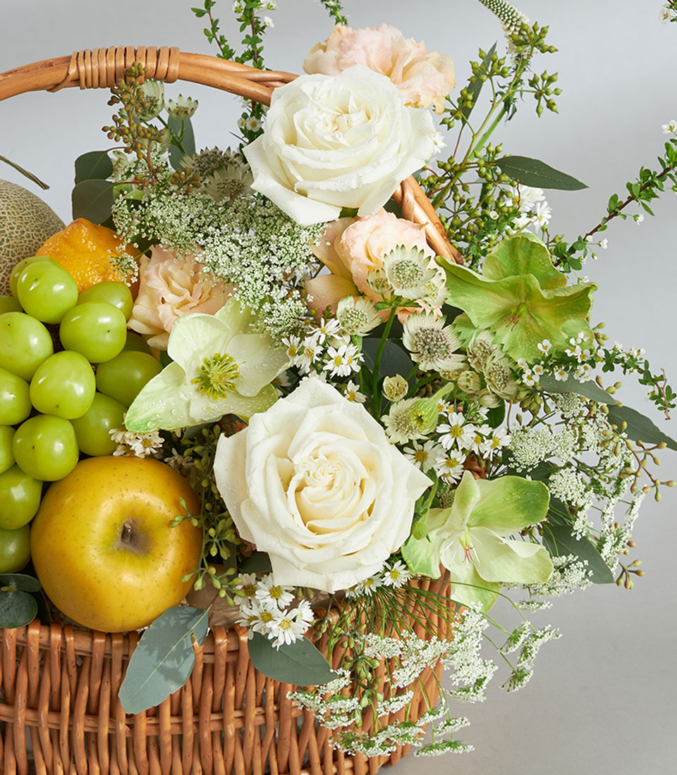 Premium green and white fruit basket with imported grapes, melon, roses, and fresh florals in woven wicker
