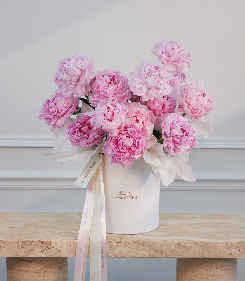Luxurious pink peonies in a white ceramic vase with "Flowers by Basketeer" branding, tied with blush ribbons on a marble surface.