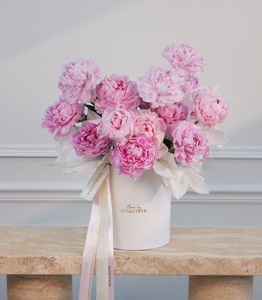 Luxurious pink peonies in a white ceramic vase with "Flowers by Basketeer" branding, tied with blush ribbons on a marble surface.
