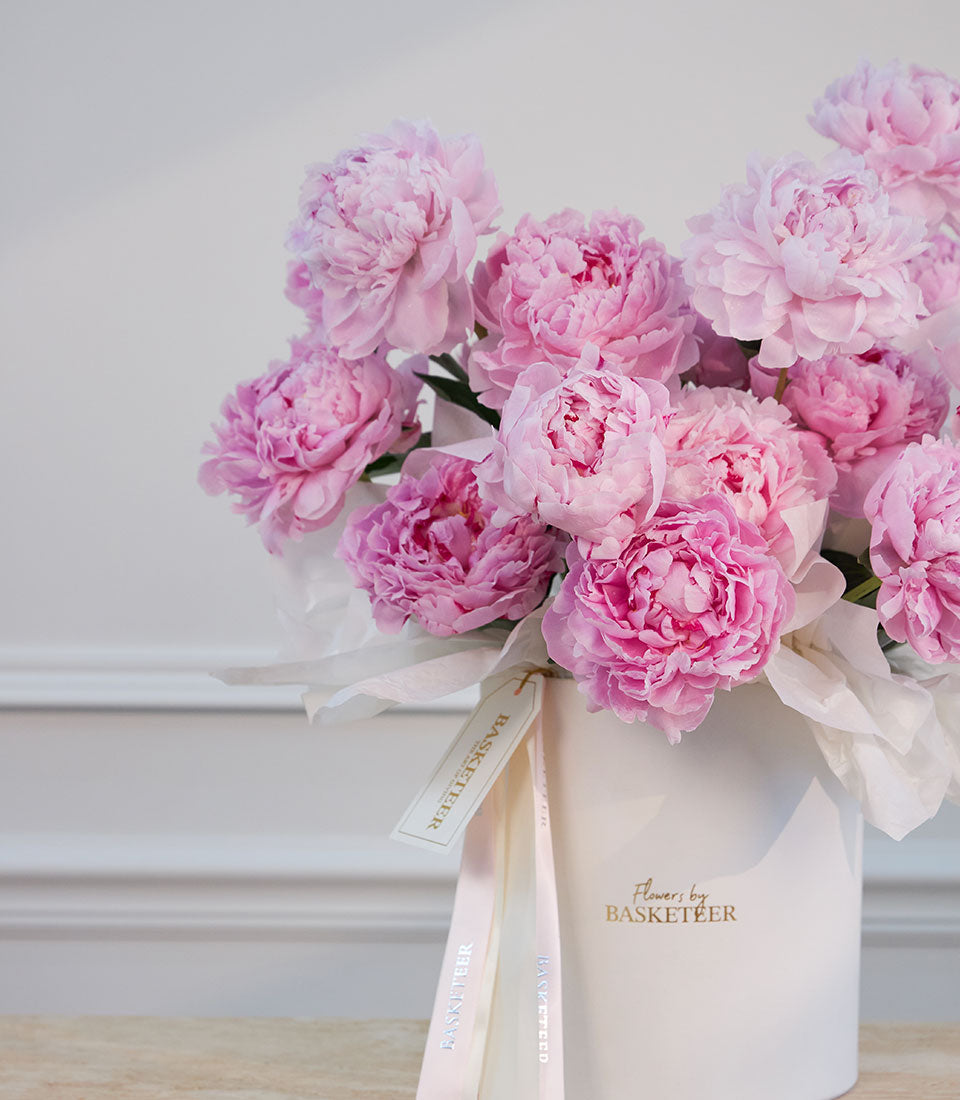 Pink peonies in a white box with cream ribbon, branded "Flowers by Basketeer" against a light gray wall.