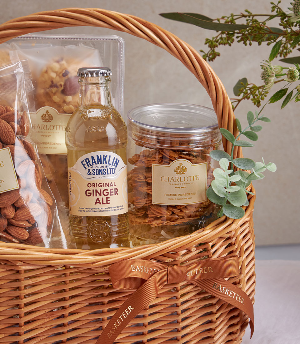 Light brown wicker basket filled with gourmet almonds, cookies, ginger ale, and golden snacks with gold-toned labels and