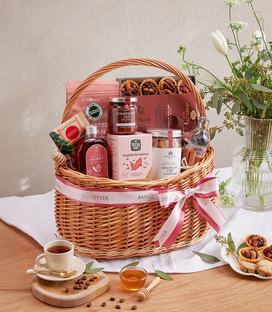 Wicker gift basket filled with gourmet coffee, tea, honey, jam, and artisan cookies with pink ribbon bow