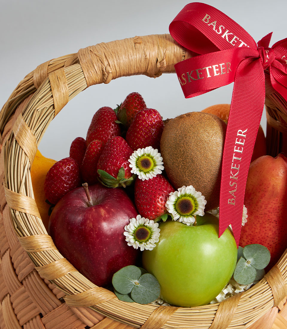 Woven wicker fruit basket with strawberries, apples, pear, kiwi, and fresh flowers tied with red grosgrain ribbon