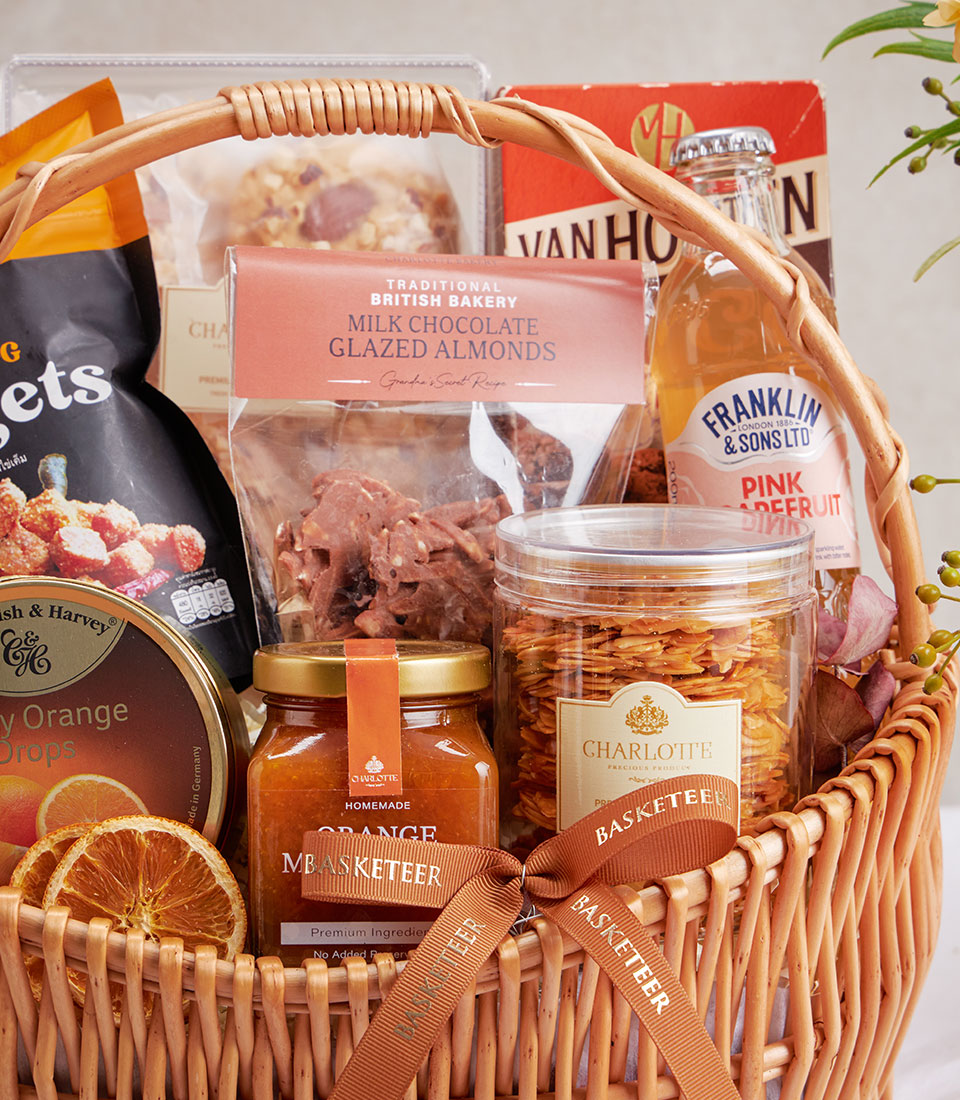Wicker gift basket with almond cookies, dark chocolates, orange drops tin, and gourmet treats with pink grapefruit beverage