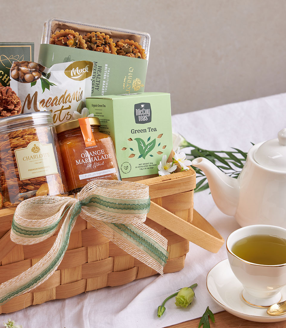 Woven gift basket with chocolate chip cookies, macadamia nuts, green tea, and marmalade on white tablecloth