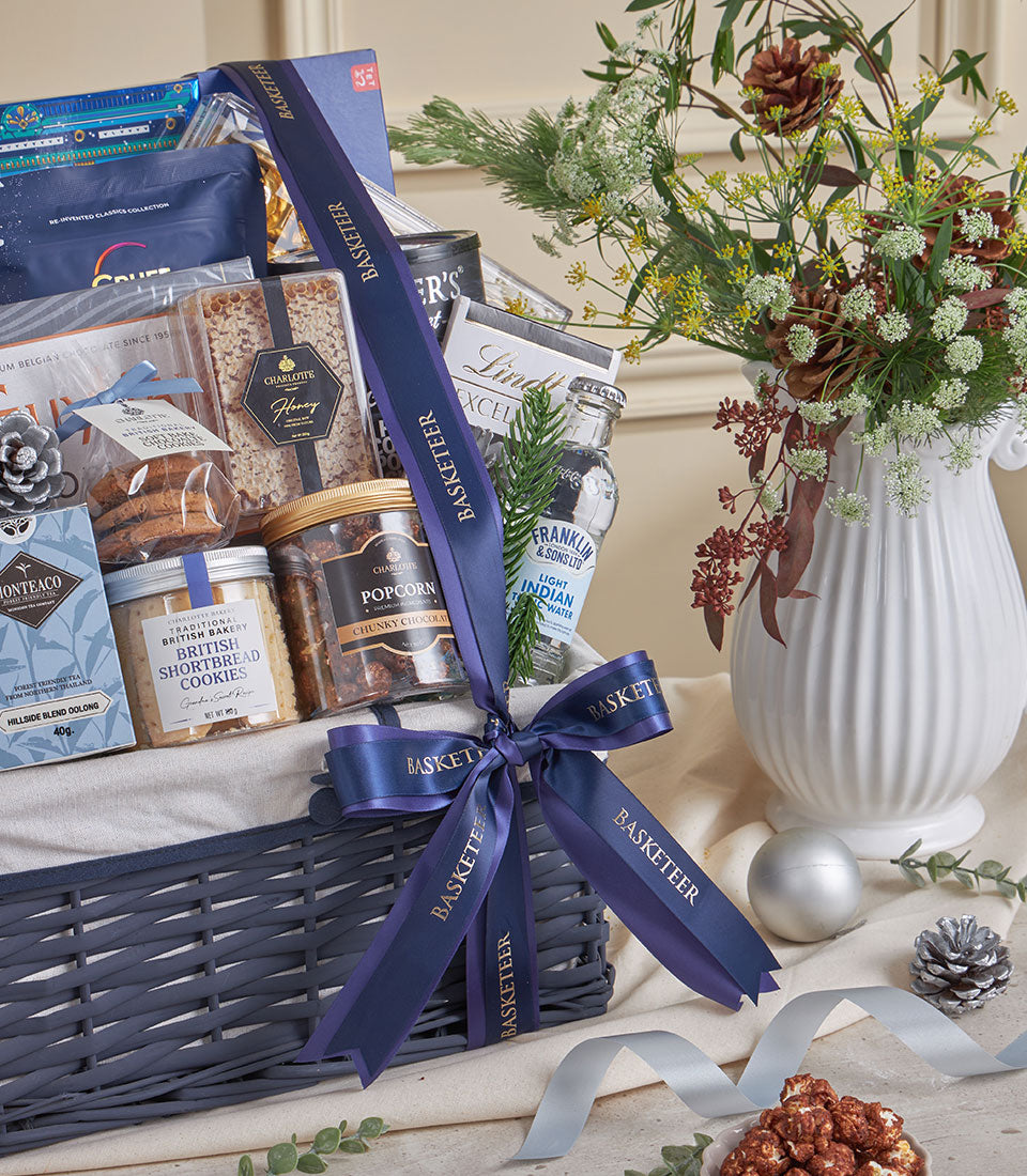 Blue woven gift basket with gourmet treats, shortbread, tea, and tonic water tied with satin ribbon for New Year celebrations
