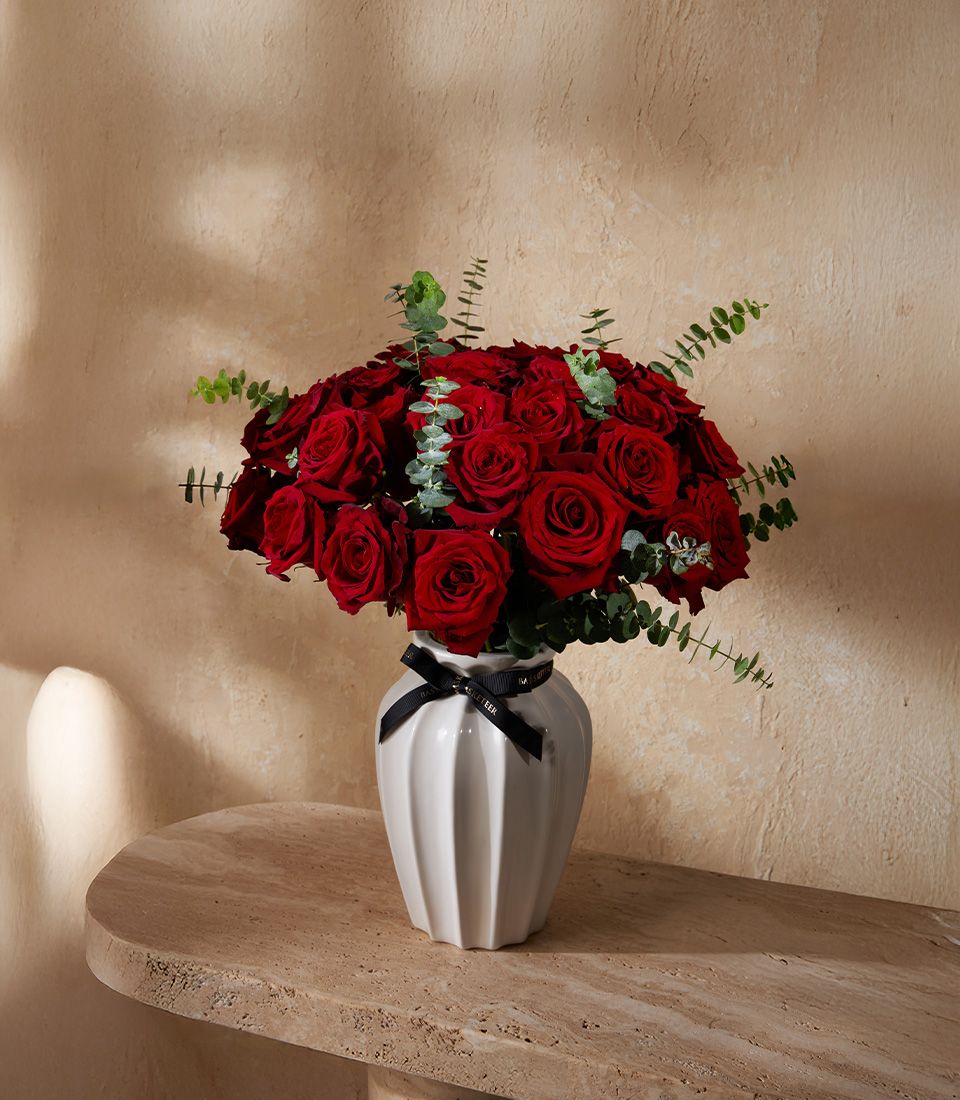 Vibrant scarlet rose bouquet in white fluted ceramic vase with black ribbon and eucalyptus greenery
