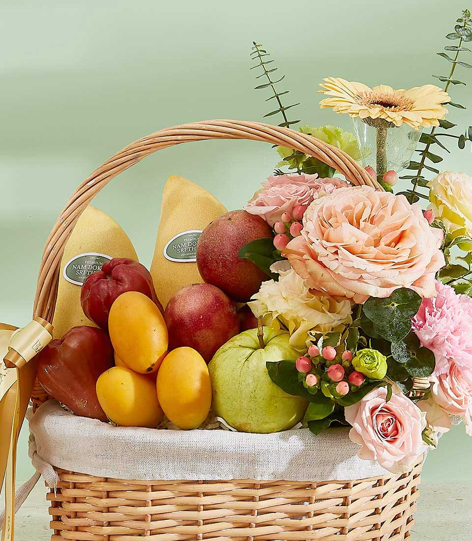 Woven wicker gift basket with premium Thai mangoes, rose apples, passion fruit, guava, and pink-peach flower arrangement