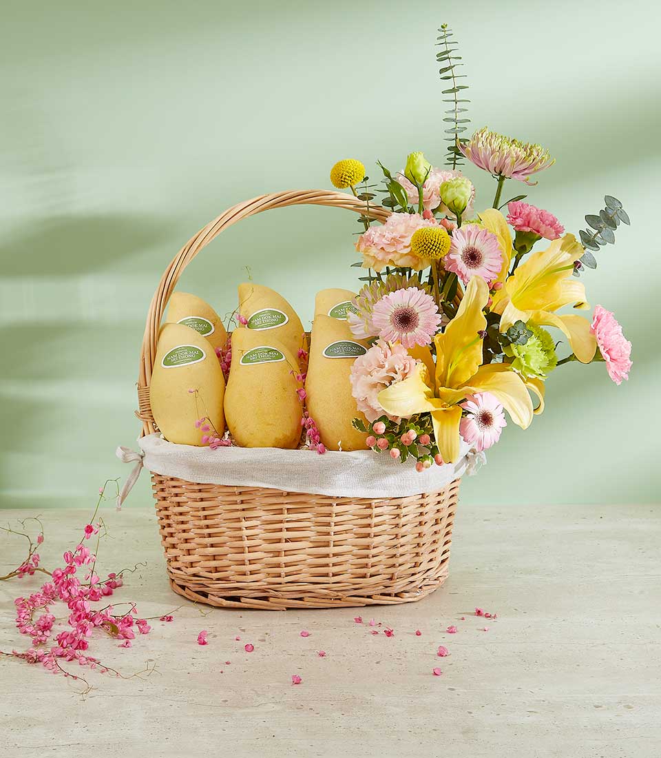 Woven basket with ripe yellow mangoes and vibrant yellow lilies, pink gerberas, and carnations on light background