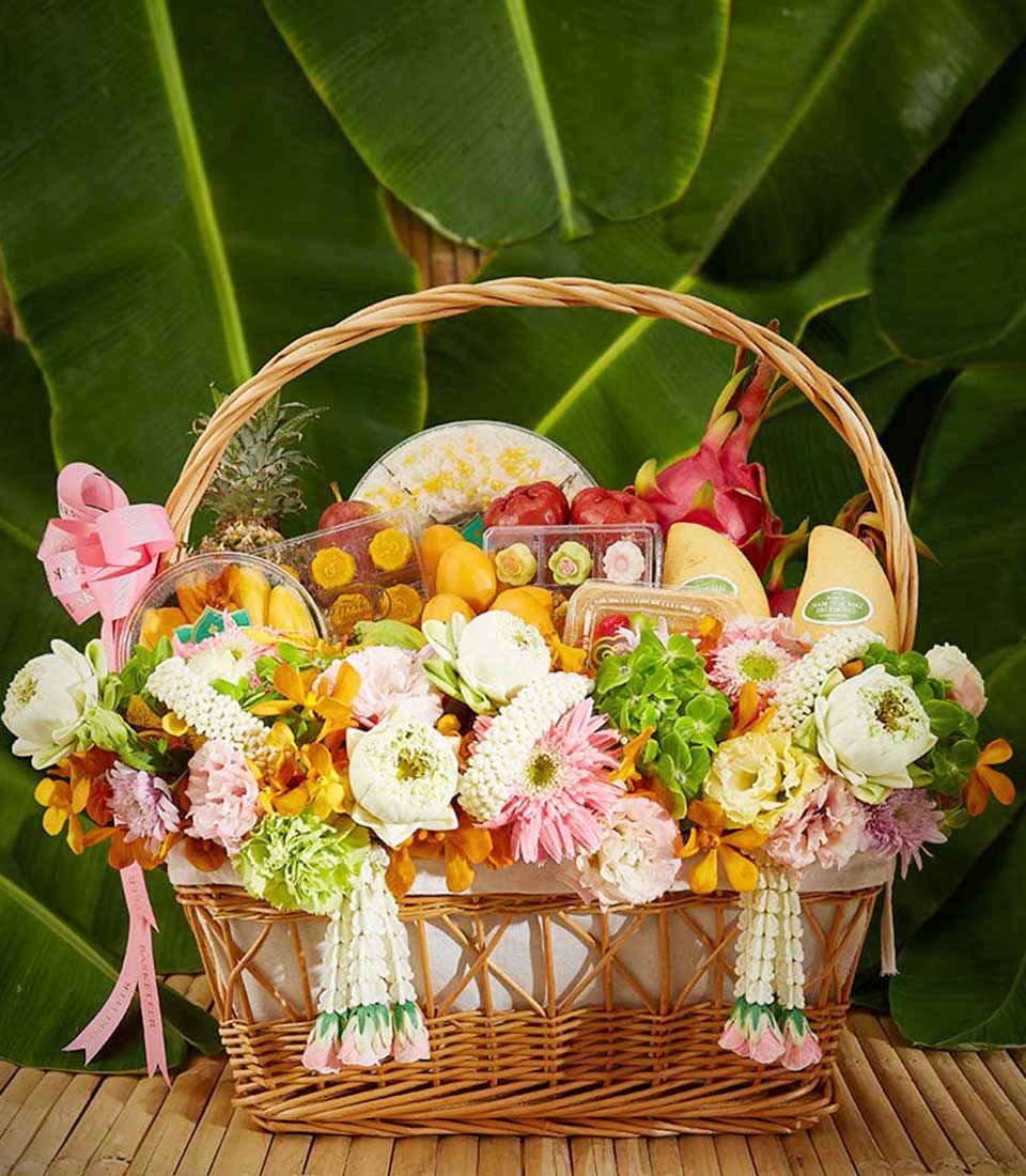 Woven wicker gift basket with fresh tropical fruits, pastries, and white, pink, yellow flowers for Thai New Year Songkran