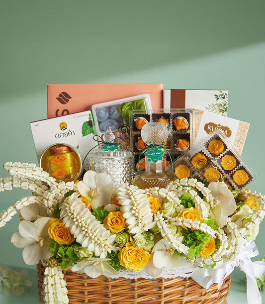 Woven wicker gift basket with white lace trim, fresh white orchids, yellow roses, jasmine garlands, and gourmet treats for