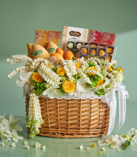 Woven wicker gift basket with white and yellow orchids, roses, Thai mangoes, coconut snacks, and traditional desserts for