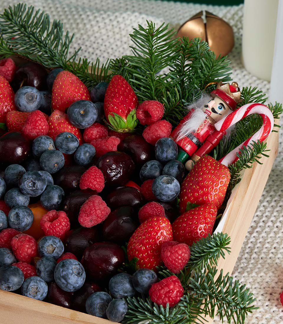 Festive wooden tray with fresh strawberries, blueberries, raspberries, cherries, pine sprigs, and holiday decorations