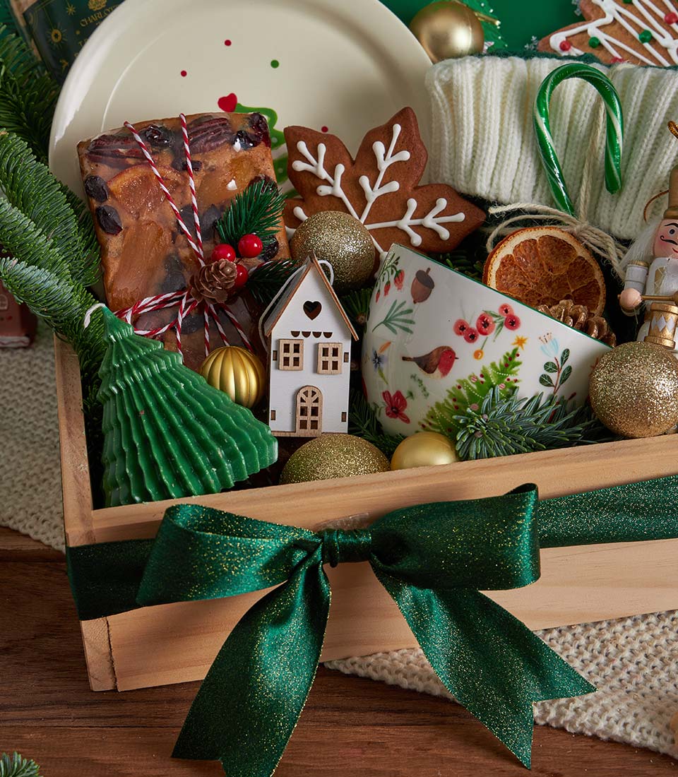 Festive wooden Christmas gift basket with fruitcake, gingerbread cookies, tree candle, and gold ornaments tied with dark