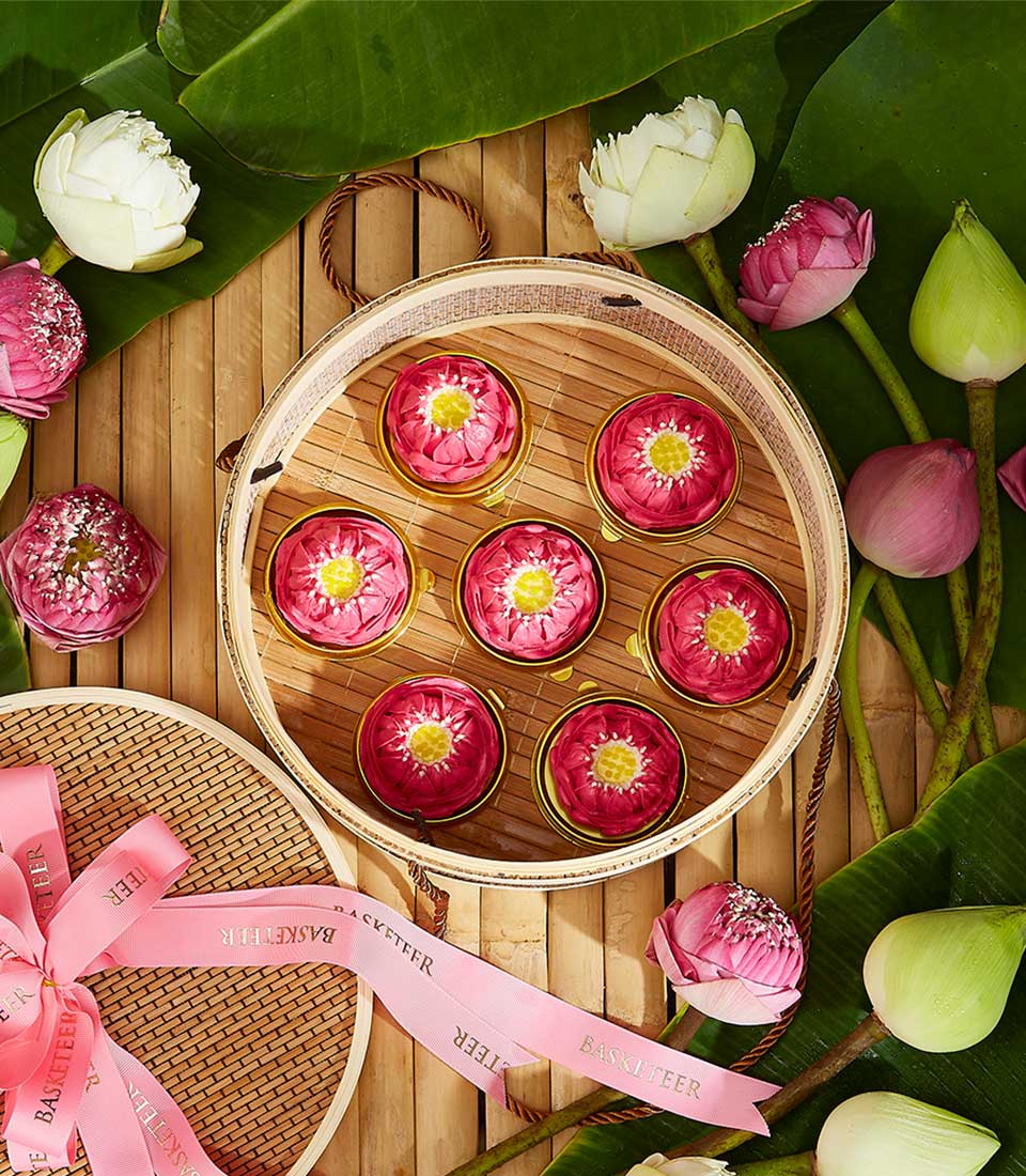 Lotus-shaped Thai dessert gift set with seven pink and gold confections on bamboo tray, surrounded by fresh white and pink