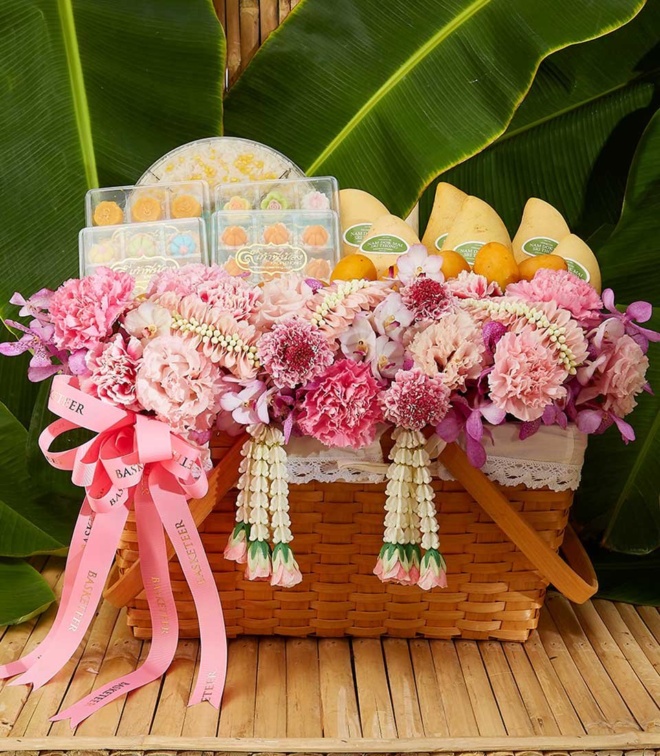 Woven wicker gift basket with pink carnations, white orchids, Thai flower garlands, Khanom Luk Chup desserts, and fresh