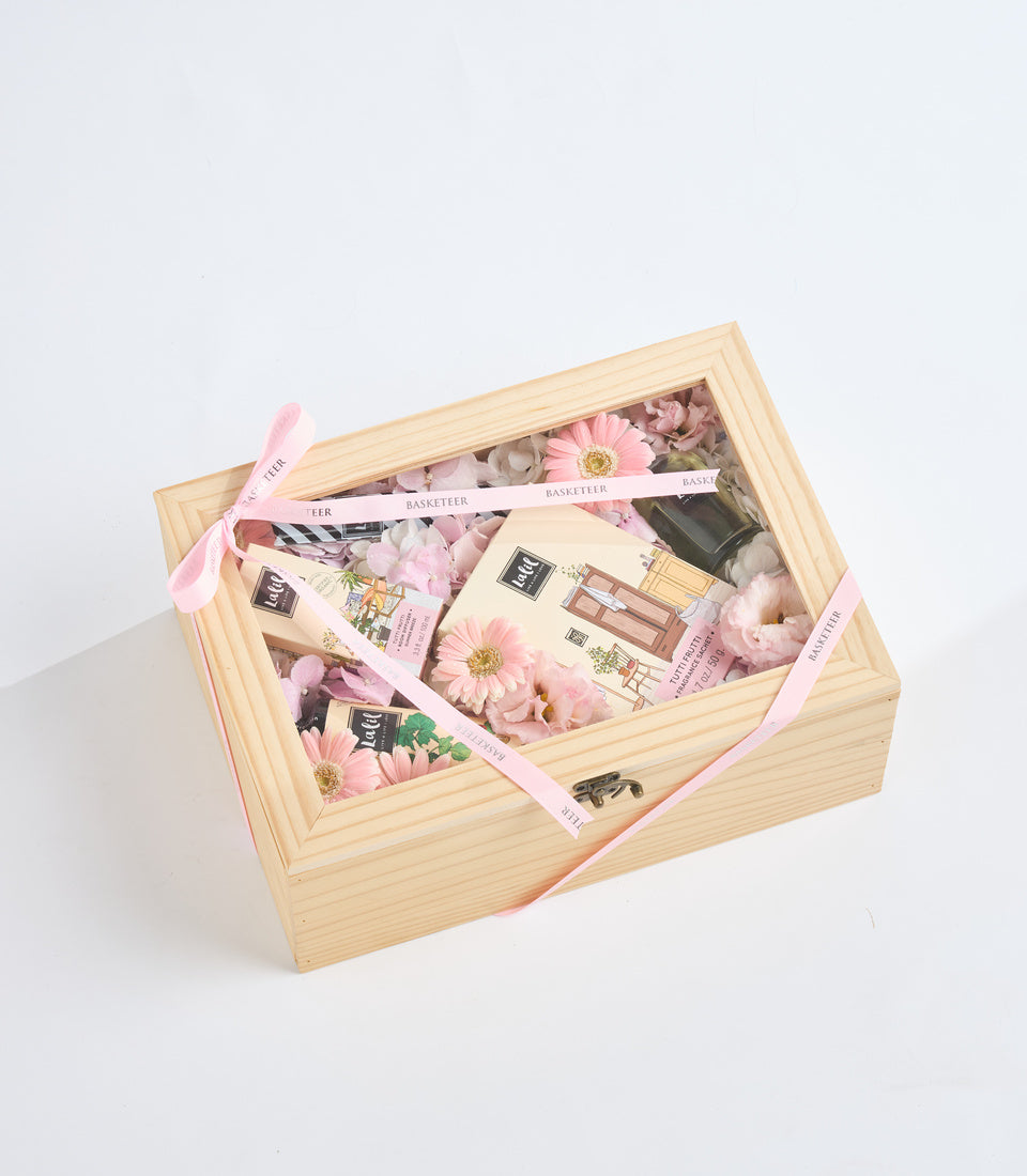 Wooden gift box filled with pink flowers, beauty products, and cosmetics, tied with pink "Basketeer" ribbon on white background.