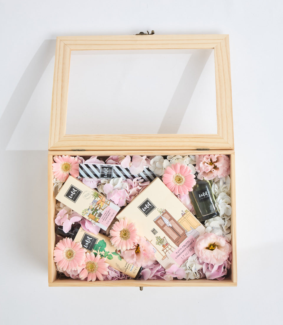Wooden gift box with glass lid containing pink flowers, beauty products, striped ribbon, and white filler, displayed open against white background.
