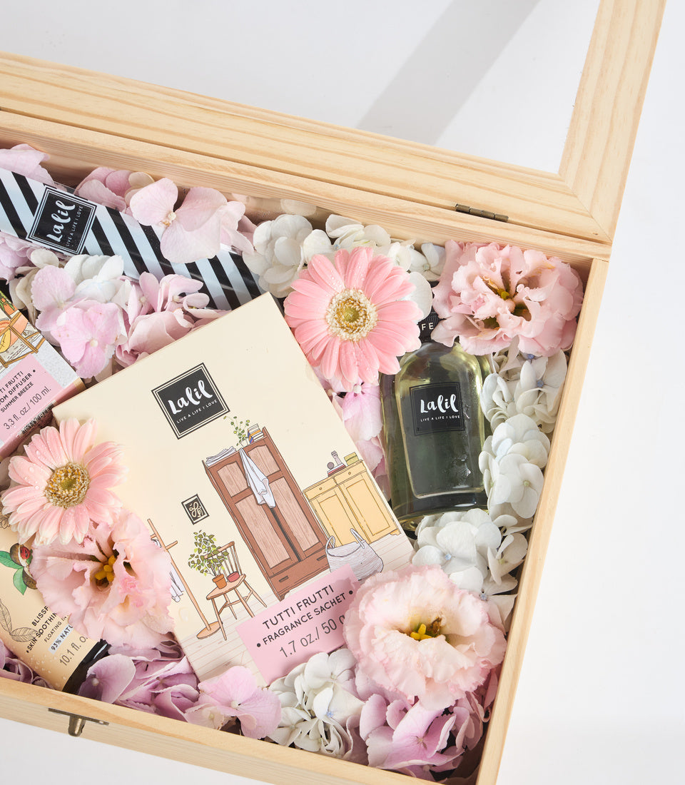 Luxury gift box with wooden frame containing pink and white flowers, Lalil beauty products, fragrance sachet, and black striped ribbon.