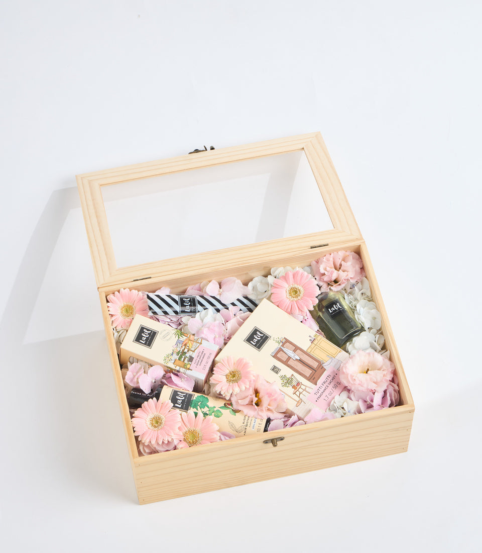 Wooden gift box with glass lid containing beauty products, pink gerbera daisies, white flowers, striped ribbon, and cosmetic items arranged on white filler.