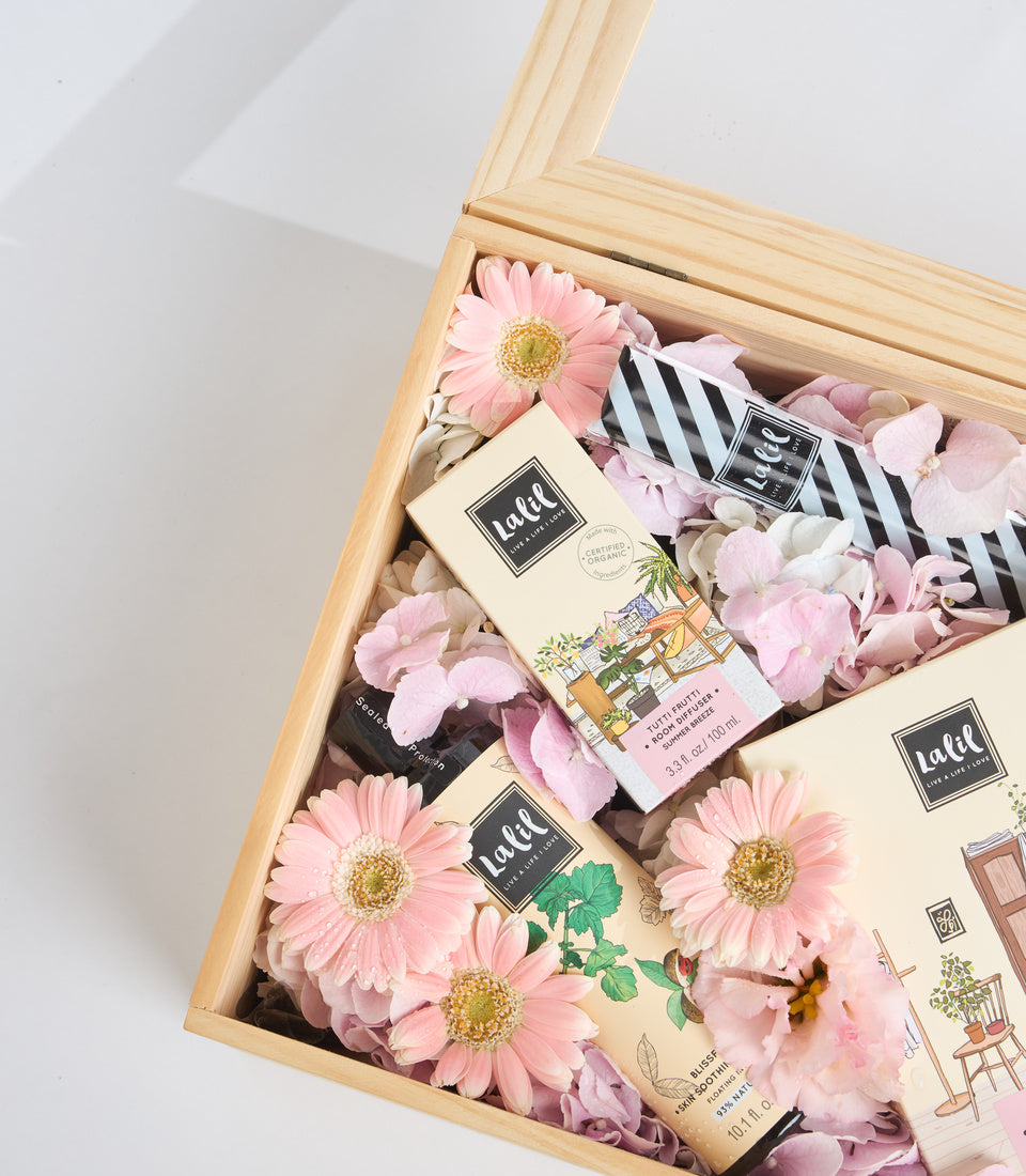 Wooden box gift set with pink gerbera daisies, Ladd skincare products, black striped ribbon, and dried flower filler on white background.
