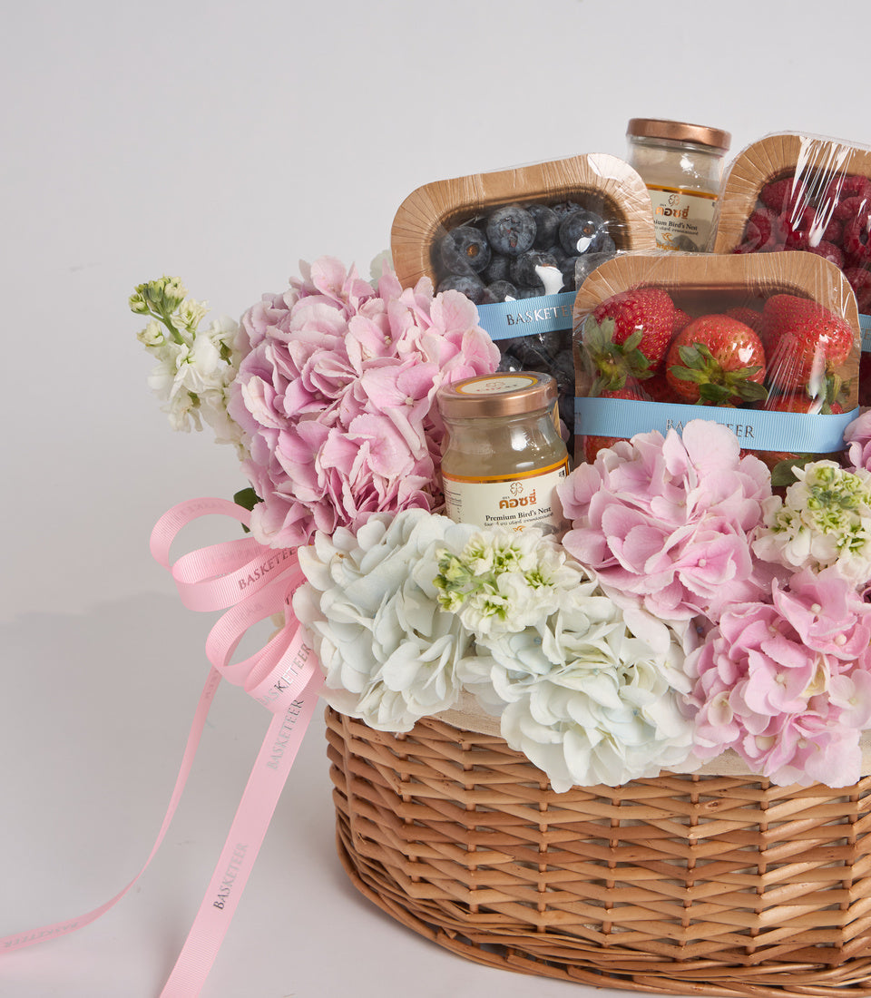 Luxury gift basket with pink and white peonies, fresh berries, gourmet treats, and beauty products in a woven wicker basket with pink ribbon.