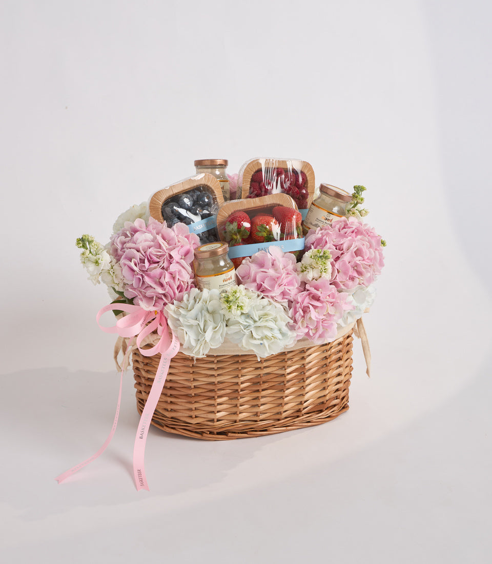 Wicker basket filled with pink and white carnations, gourmet treats including fresh strawberries, jams, and packaged goods, tied with pink ribbon.