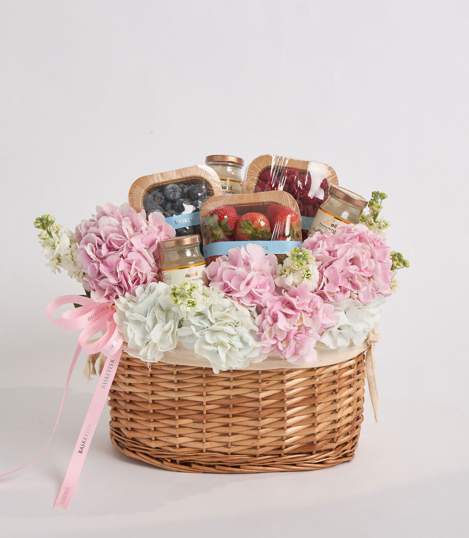 Wicker basket filled with pink and white flowers, gourmet jars of preserves, and jams tied with pink ribbon on white background.
