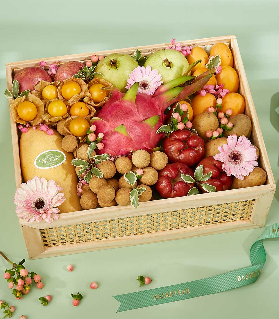 Premium Thai tropical fruits in woven wooden crate with mango, pomegranate, dragon fruit, and fresh flowers