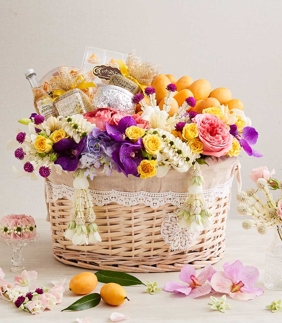 Woven wicker harvest basket with yellow and pink roses, purple orchids, tropical mangoes, and gold-wrapped gifts for