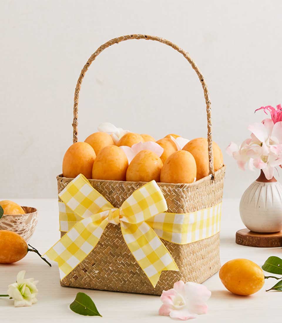 Woven straw basket filled with golden Marian plums tied with yellow gingham ribbon, surrounded by fresh tropical fruits and