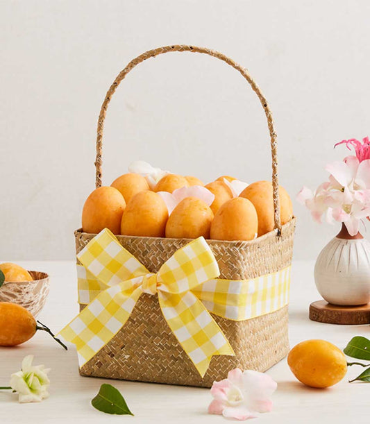 Woven straw basket filled with golden Marian plums tied with yellow gingham ribbon, surrounded by fresh tropical fruits and