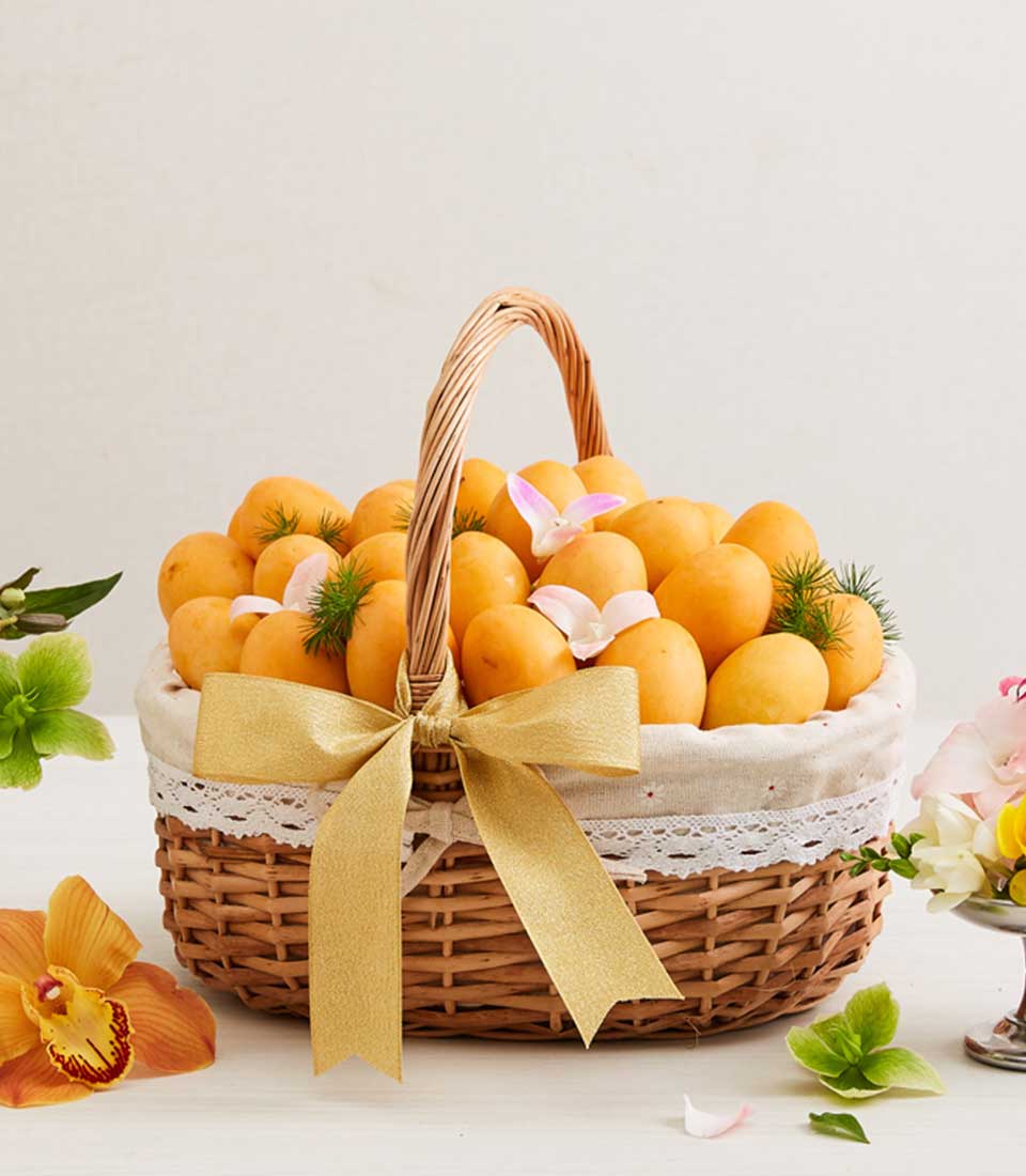 Premium wicker hamper overflowing with ripe golden mangoes, white lace trim, gold ribbon bow, and fresh orchid blooms