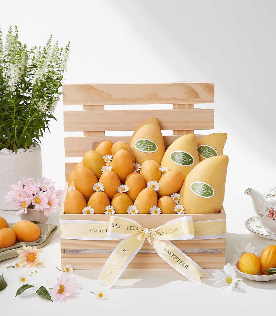 Light wooden crate of premium Nam Dok Mai and Mayongchid mangoes with yellow ribbon, fresh flowers, and artisan gift