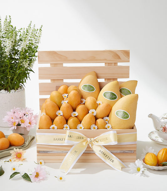 Light wooden crate of premium Nam Dok Mai and Mayongchid mangoes with yellow ribbon, fresh flowers, and artisan gift