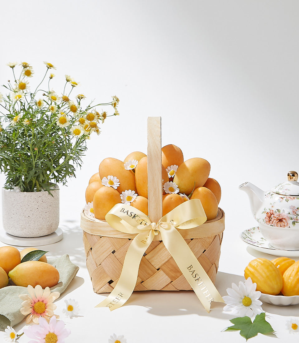 Woven gift basket filled with ripe golden mangoes and white daisies, tied with Basketeer satin ribbon