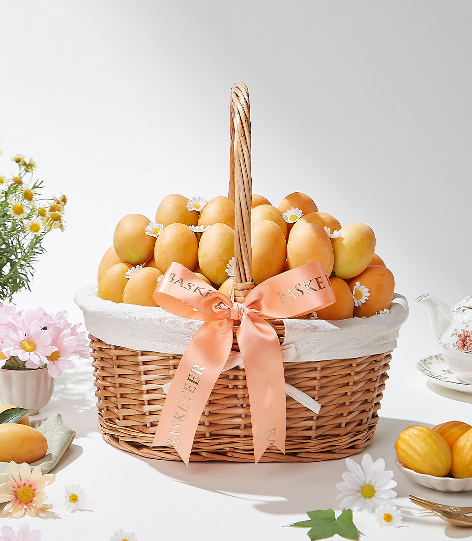 Wicker gift basket overflowing with golden mangoes, white daisies, and peach satin ribbon bow