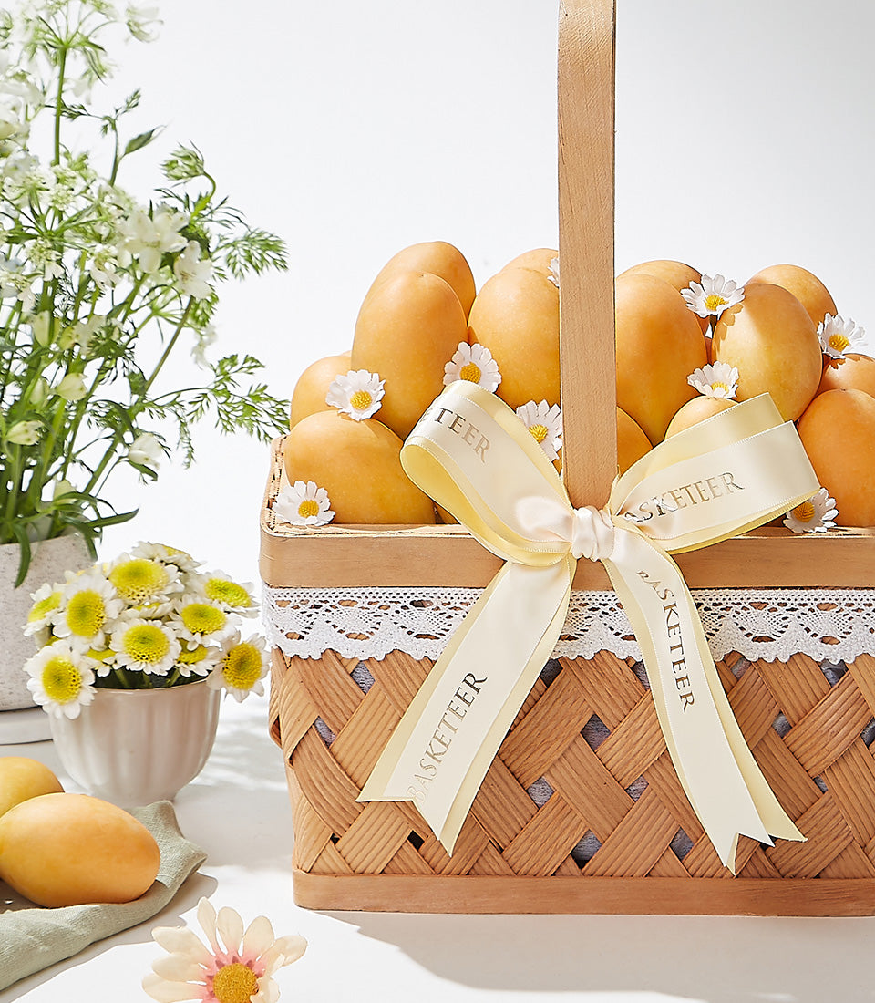 Luxury woven gift basket overflowing with ripe golden mangoes, cream ribbon bow, and white daisy accents