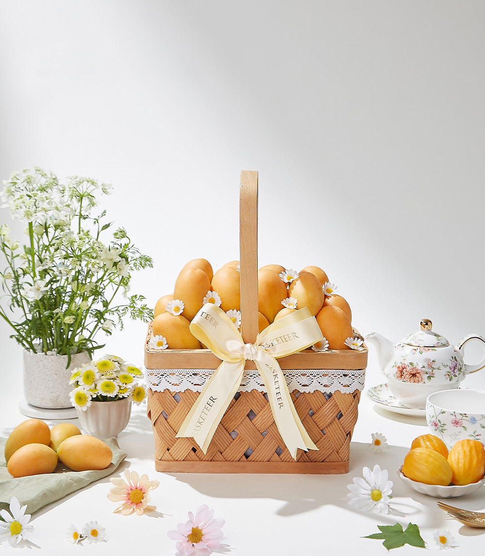 Woven gift basket overflowing with golden tropical fruits, white daisies, cream ribbon bow, and premium artisan accessories