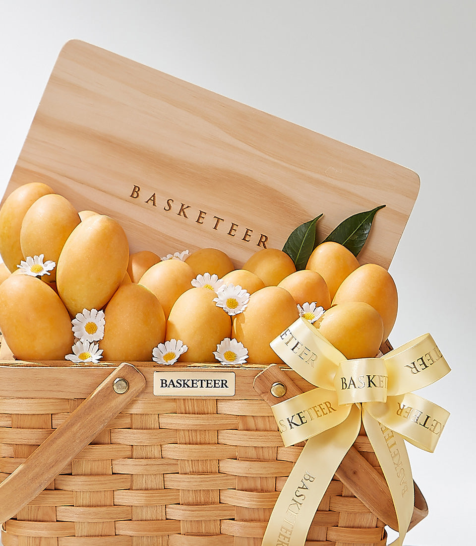 Woven picnic basket filled with Thai yellow mangoes, white daisies, and green leaves, tied with light yellow ribbon
