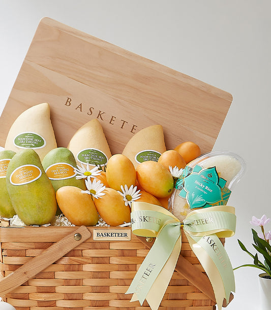 Golden woven gift basket with Thai mangoes, sticky rice, and white flowers, tied with cream and green ribbon