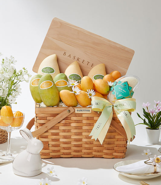 Woven basket filled with Thai mangoes, sticky rice, and light green ribbon bow, surrounded by white daisies and madeleine
