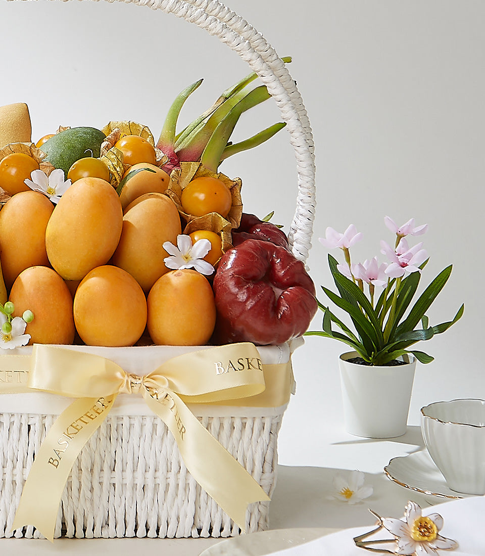 White woven gift basket filled with fresh tropical mangoes, goldenberries, and rose apples, tied with gold satin ribbon