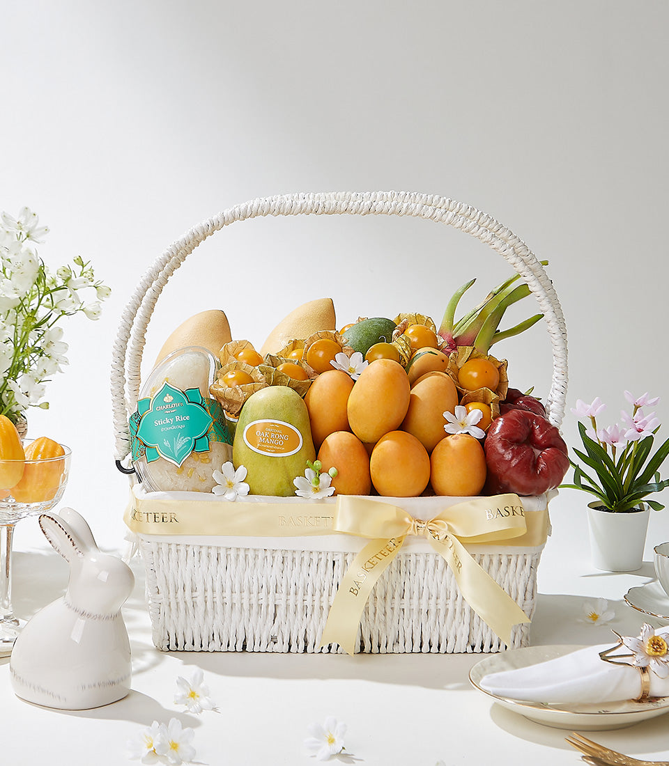 Luxury white wicker fruit basket with golden mangoes, physalis, and Thai tropical fruits tied with gold satin ribbon