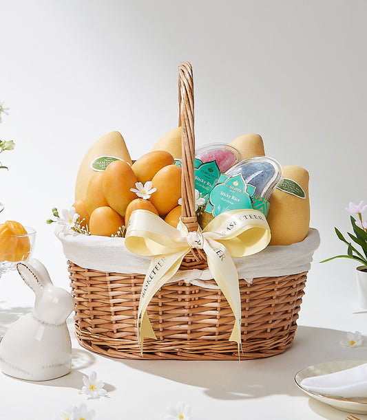 Woven wicker basket overflowing with golden Thai mangoes, sticky rice, and white flowers tied with cream Basketeer ribbon
