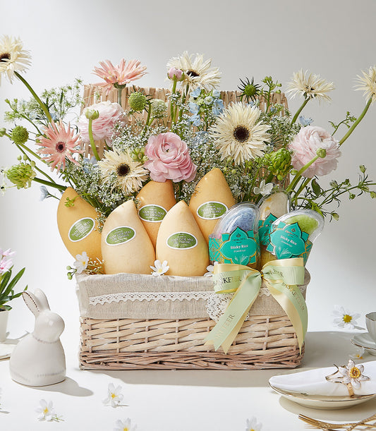 Woven gift basket with Thai Nam Doc Mai mangoes, sticky rice, and white pink blue flower arrangement