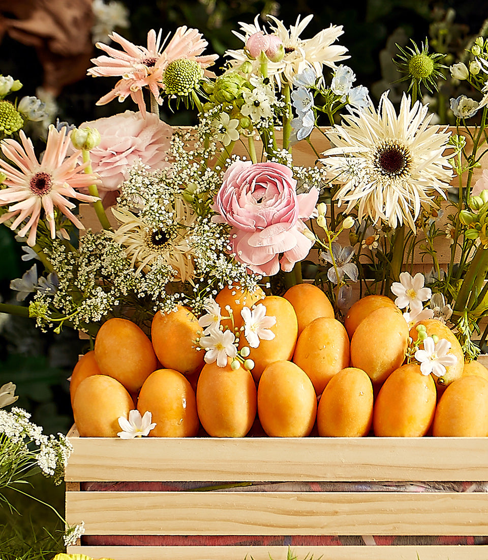 Wooden crate of golden Thai tropical fruits with pink and white gerbera daisies, ranunculus, and baby's breath