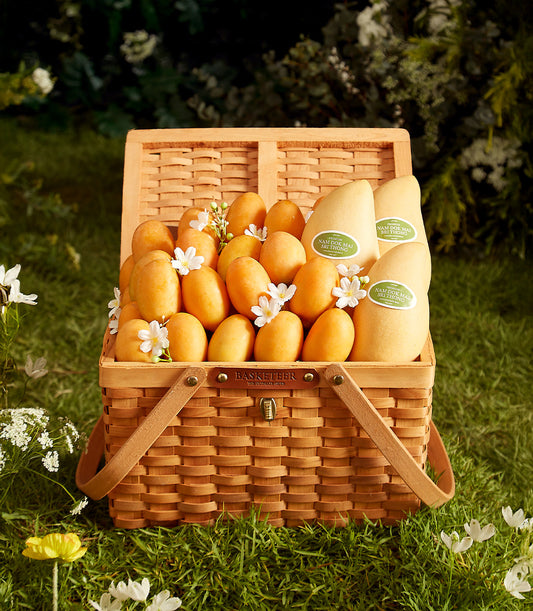 Woven picnic basket filled with fresh Thai mangoes and white flowers on green grass
