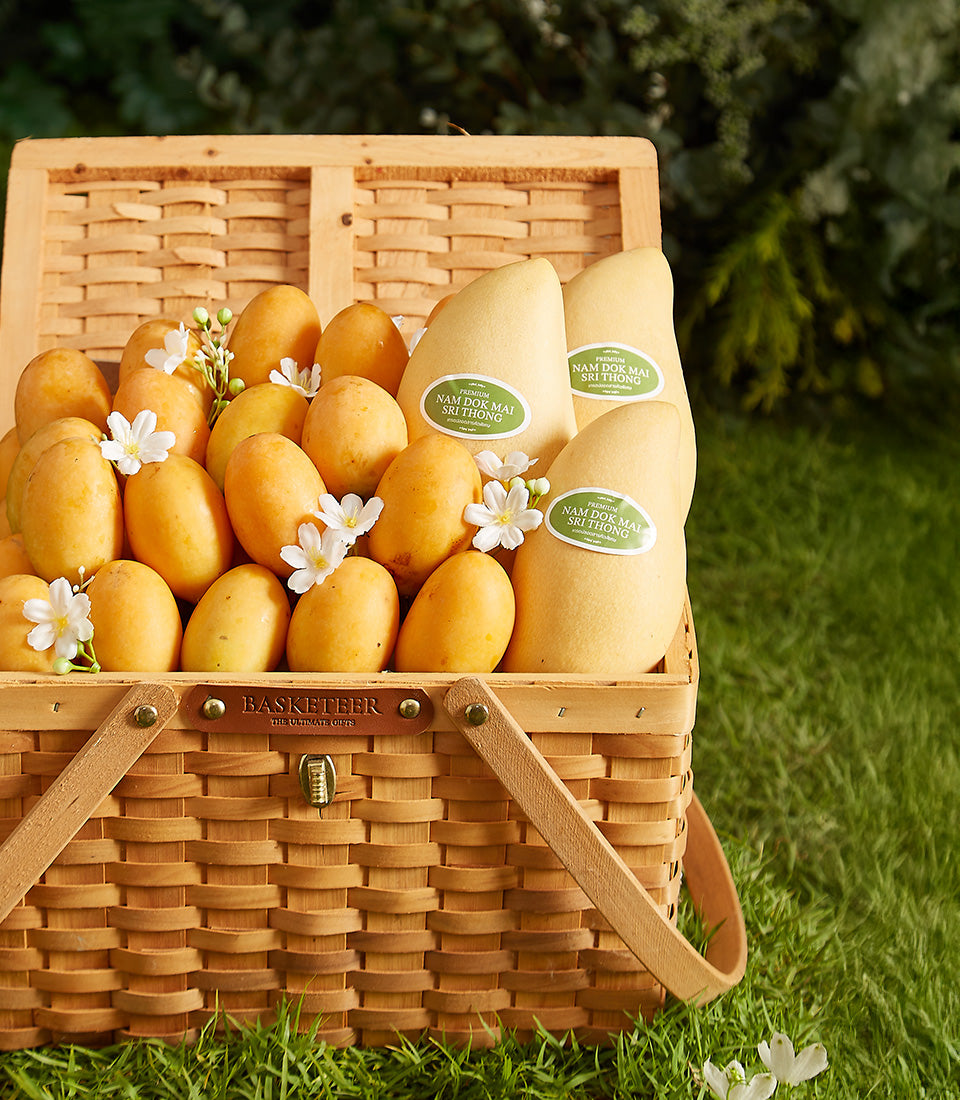 Woven picnic basket filled with Thai Nam Dok Mai mangoes and white flowers, luxury tropical fruit gift hamper